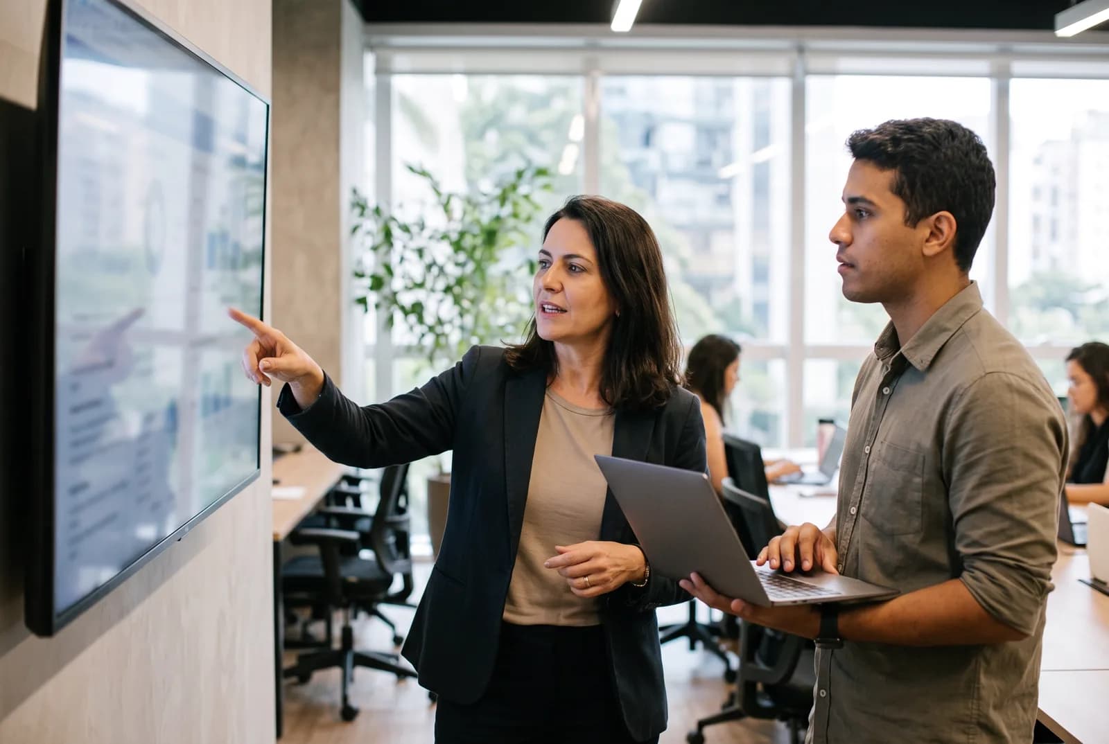 Gestora comercial em pé, apontando para um monitor grande na parede enquanto conversa com um colega que segura um notebook.